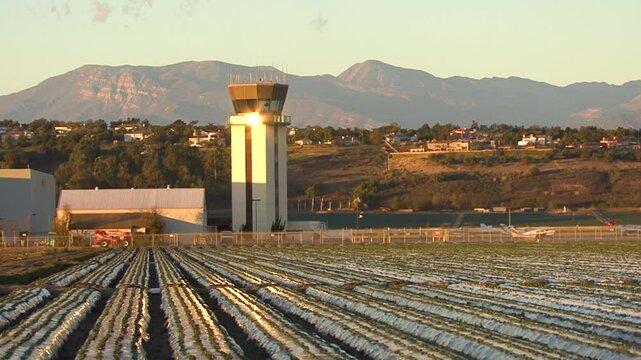 An airport control tower looks out over runways and the adjacent strawberry farm, in Camarillo, California.  Aircraft taxi about against a background of hills and the high mountains.