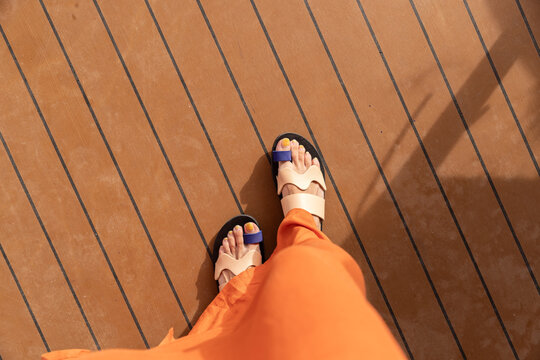 Female in flowy orange maxi dress looking down at her feet with stylish artistic sandals on a wood floor. 