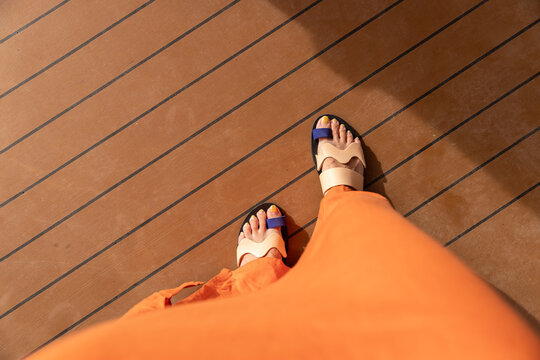 Female in flowy orange maxi dress looking down at her feet with stylish artistic sandals on a wood floor. 