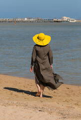 A woman in a long dress and a yellow hat walks along the coastline of the Red Sea. Everyday life of tourists. Hurghada, Governorate - Red Sea, Egypt.