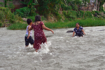 Fototapeta premium A mother sits with her legs outstretched in the stream, while two children play around her, walking and enjoying the refreshing water.