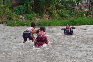 The children bend down playing with water, splashing joyfully while the mother watches calmly from...