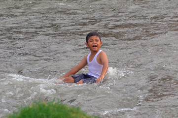 Fototapeta premium A young boy enjoys the river alone, stretching his legs while sitting calmly in the refreshing water flow, surrounded by the peaceful grassy riverbank.
