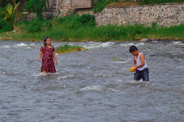 A boy successfully picks up a drifting coconut husk from the flowing river as a girl watches, both...
