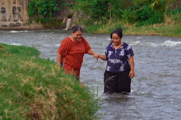 On the grassy riverbank, one woman extends her hand to assist another in stepping carefully into...
