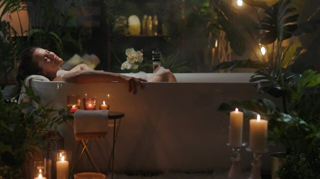 A woman relaxes in a bubble bath with candles and plants in a dimly lit bathroom. A calming and luxurious mood suggestive of self-care and relaxation.