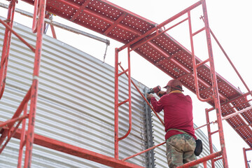 The worker is standing on the scaffold, using a cordless impact wrench to tighten bolts on the metal sheet tank wall.
