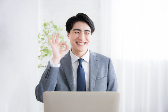 デスクで座るスマホで通話するパソコンを使うビジネスマン カメラ目線の笑顔のオッケー