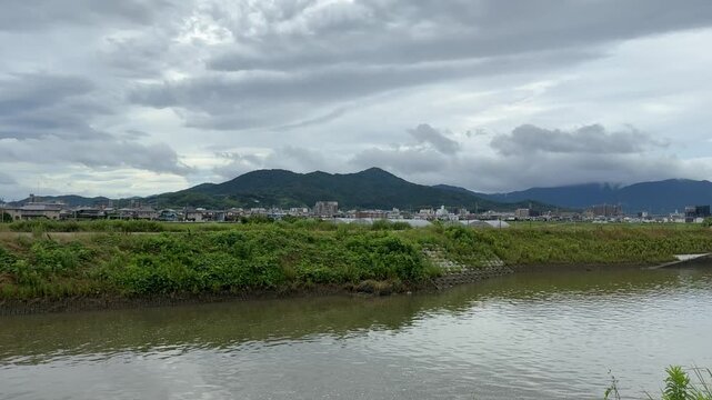 A real-time panning footage of rural Japan featuring a calm river, distant mountains, and green fields under a cloudy sky. Captured with original ambient sounds including flowing water, wind, and bird