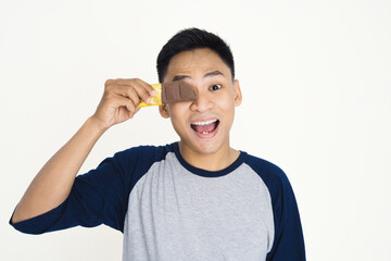 Young man holding golden-wrapped chocolate bar over his eye, expressing playful enthusiasm. Ideal image for themes of happiness, fun, sweets, and youthful enjoyment.