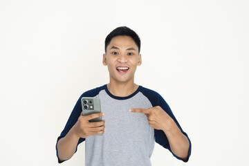 A cheerful young man wearing a casual shirt, joyfully holding a smartphone and pointing to it, expressing amazement.