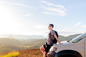 Traveler leaning on his car at sunset, looking at the mountains. A moment of freedom, inspiration, and connection with nature during a road trip.