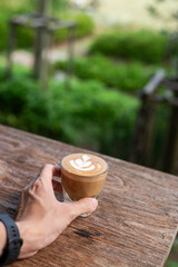 Hand holding a glass of latte on a wooden table with a blurred background.