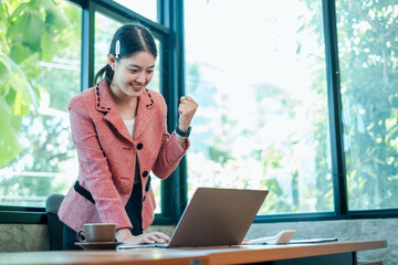 A joyful businesswoman in a pink blazer celebrates success while working on a laptop in a bright,...