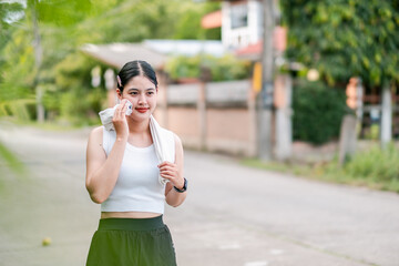 A young woman wipes sweat with a towel after a workout, standing on a suburban street with greenery...