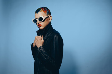 young woman with colorful hair and sunglasses wearing a black leather jacket against a blue...