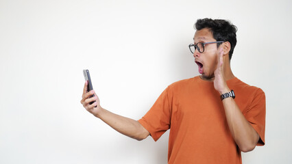 A man in an orange shirt is holding a cell phone up to his face, looking shocked
