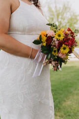 Bride holding a vibrant bouquet of sunflowers, red roses, and pink blooms, wearing a white...