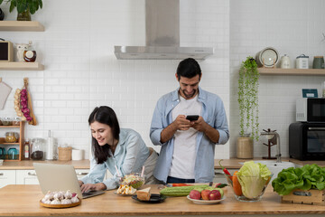 Couple using laptop and smartphone in kitchen while preparing food, enjoying modern digital lifestyle at home
