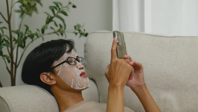Asian man relaxing with face mask while using smartphone. - Powered by Adobe