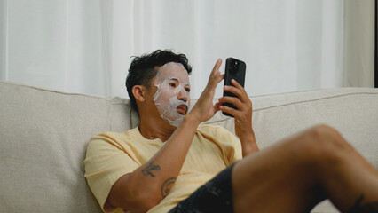 Asian man relaxing with sheet mask while using smartphone.