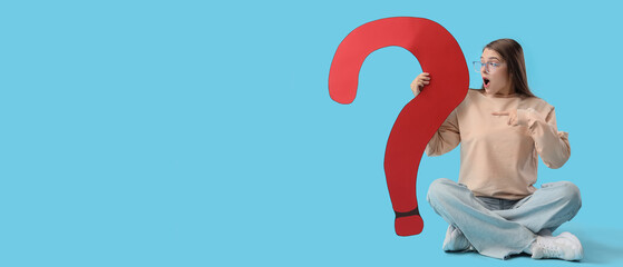 Shocked young woman pointing at big paper question mark on blue background