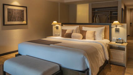 A double bed with white sheets and a beige bedspread. Pillows at the headboard. The couch. The phone is on the bedside table. The lamps are lit. The interior of a bedroom in a hotel. The suite.