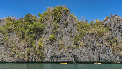 Tourist canoes sail along a sheer cliff. Green tropical vegetation on steep furrowed slopes. Turquoise ocean. Blue sky, clouds. Philippines. Palawan. Big lagoon. Bacuit  Bay.