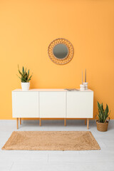 Modern white chest of drawers with houseplants and mirror near orange wall in room