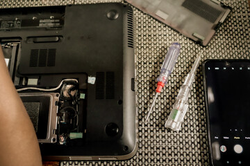 A broken laptop is under repair with a screwdriver,  a test pen and a smartphone on the table