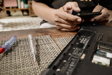Male person searching for instructions from the internet using smartphone to repair a broken laptop...
