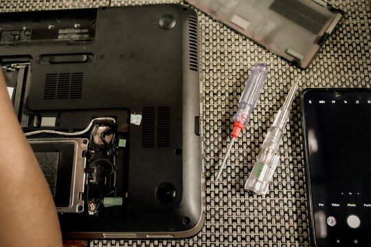 A broken laptop is under repair with a screwdriver,  a test pen and a smartphone on the table