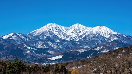 Fototapeta premium Majestic snow-capped mountain under a clear blue sky, surrounded by forested hills and vibrant natural scenery.