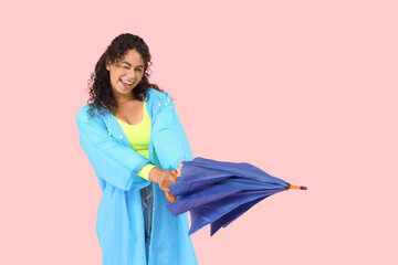Happy young African-American woman in raincoat with umbrella on pink background