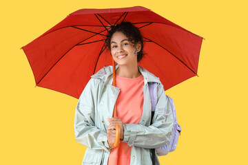 Happy young African-American woman in raincoat with umbrella on yellow background