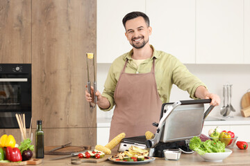 Fototapeta na wymiar Young happy man cooking fresh corn cobs on modern electric grill in kitchen