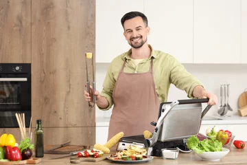 Fototapete Young happy man cooking fresh corn cobs on modern electric grill in kitchen © Pixel-Shot
