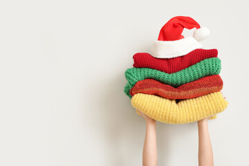 Woman holding stack of winter clothes with Santa hat near white wall