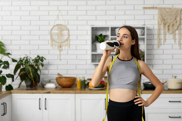 Sporty young woman with tape measure drinking water in kitchen. Diet concept