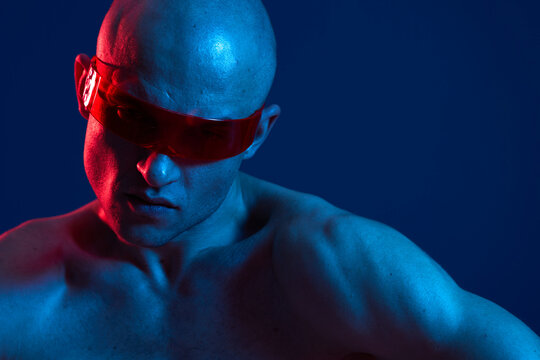 Portrait of a focused man with a muscular build, wearing red sunglasses, glowing with vibrant blue and red light, highlighting his features and strong expression.