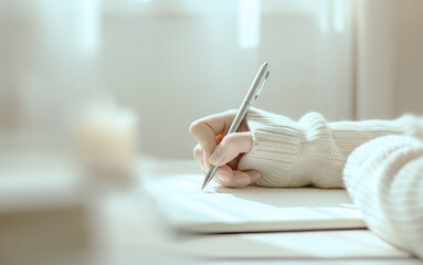 Hand in a cozy cream sweater holds a silver pen while writing on a notebook in soft natural light, creating a peaceful moment of journaling or personal reflection.