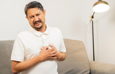 Mature Asian man sitting on sofa clutching and having chest pain cause from heart attack.