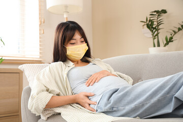 Pregnant Asian woman in yellow medical mask lying on sofa at home