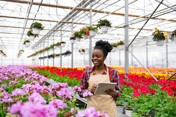 An African-American woman examines blooming flowers in a greenhouse, using a tablet to monitor plant health and growth.