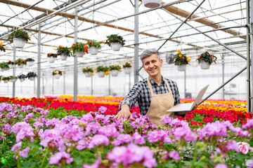 A smiling greenhouse worker examines flowers with a laptop, surrounded by vibrant blooms in a colorful floral arrangement.