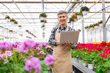 A smiling florist stands in a greenhouse, using a laptop to manage the plants.