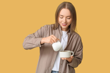 Beautiful young woman pouring fresh milk from jug into cup of hot coffee on yellow background