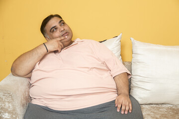 Overweight fat indian man thinking while sitting on couch at home. Plus size guy contemplate about something or day dreaming. Obesity and weight loss concept.