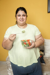 Happy Overweight fat indian woman eating healthy vegetable salad food at home. Plus size female dieting to lose weight. Obesity concept.