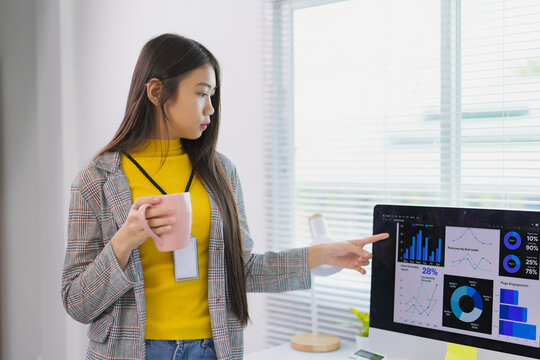 Asian businesswoman drinking coffee and pointing at computer screen showing graphs and charts of financial data, marketing analysis, business statistics and planning